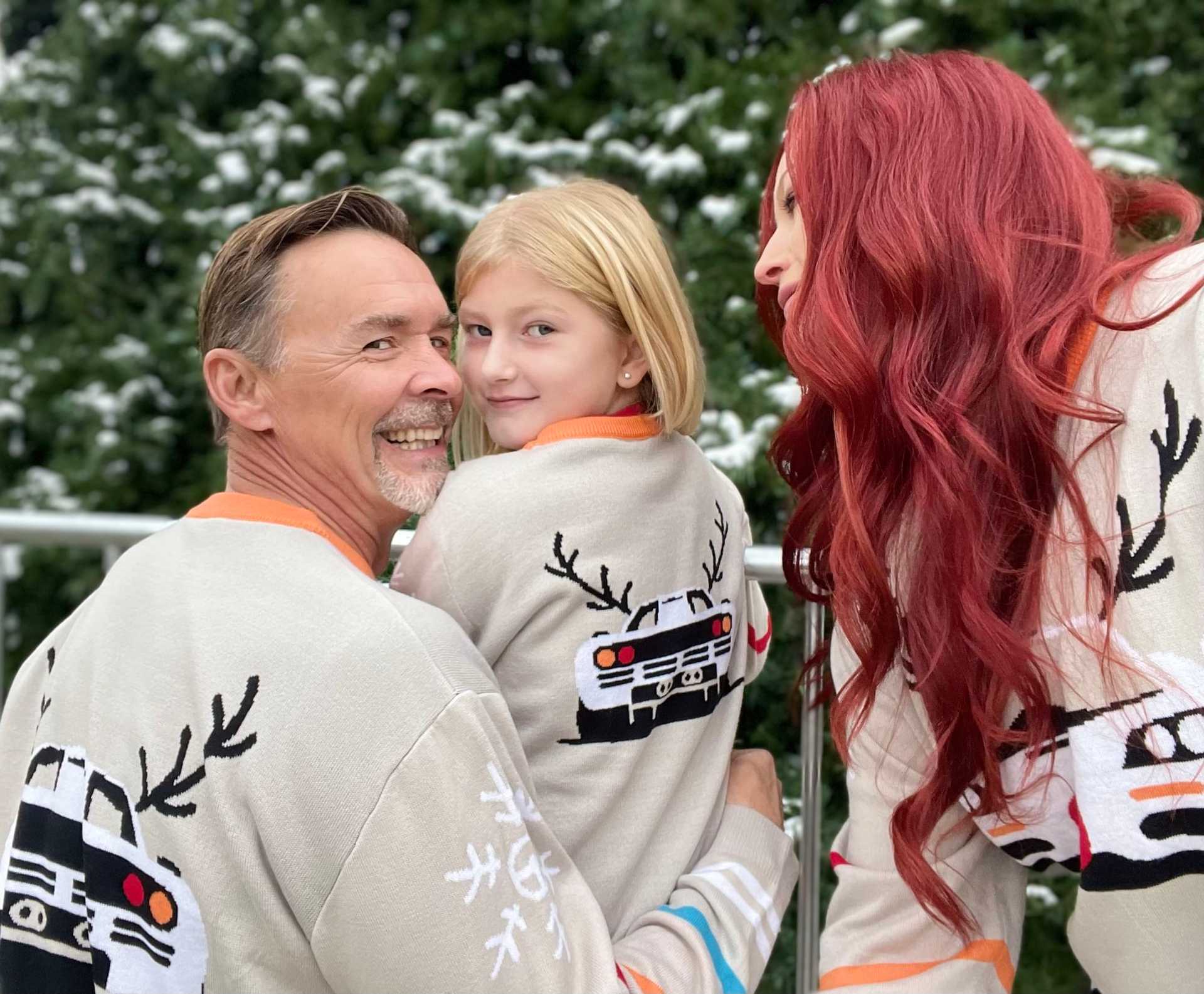 Family wearing matching reindeer sweaters, smiling in front of a snowy evergreen backdrop.