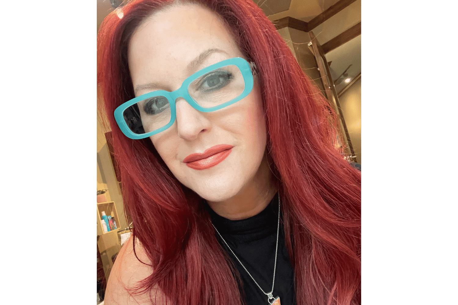 Woman with long red hair, wearing turquoise glasses, black top, and red lipstick in a well-lit room.