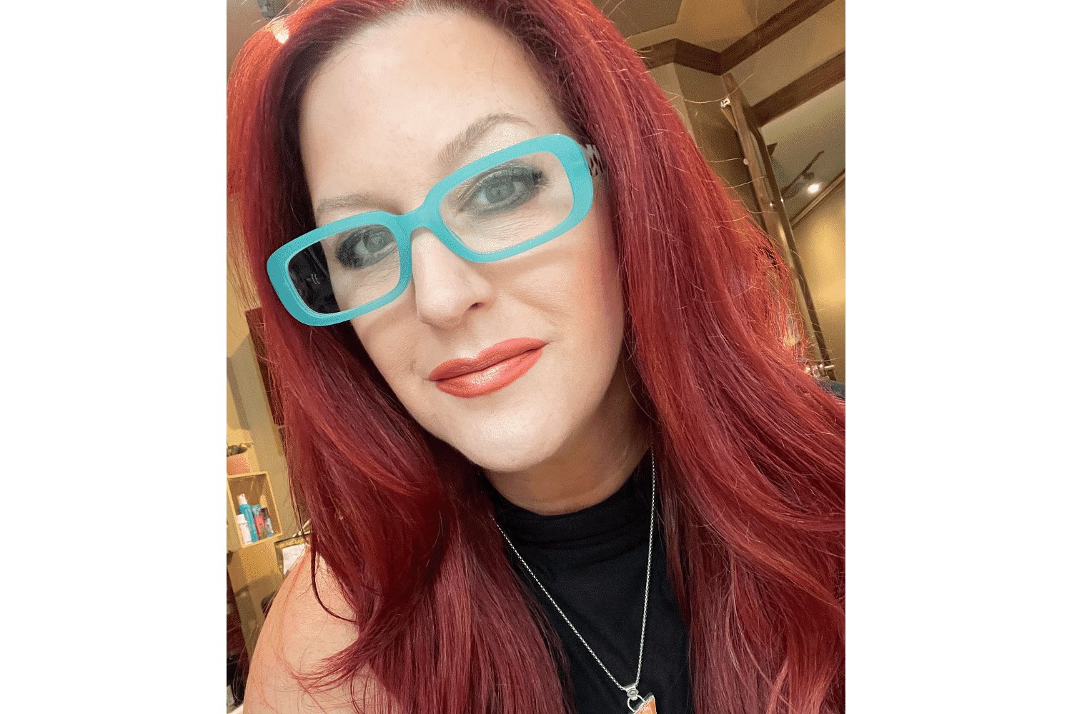 Woman with long red hair, wearing turquoise glasses, black top, and red lipstick in a well-lit room.